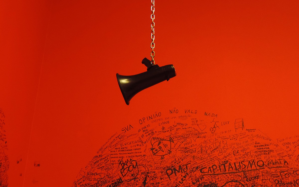 A black megaphone hanging upside down infront of a red wall covered with writing written in sharpie. The word “capitalismo” stands out on the wall