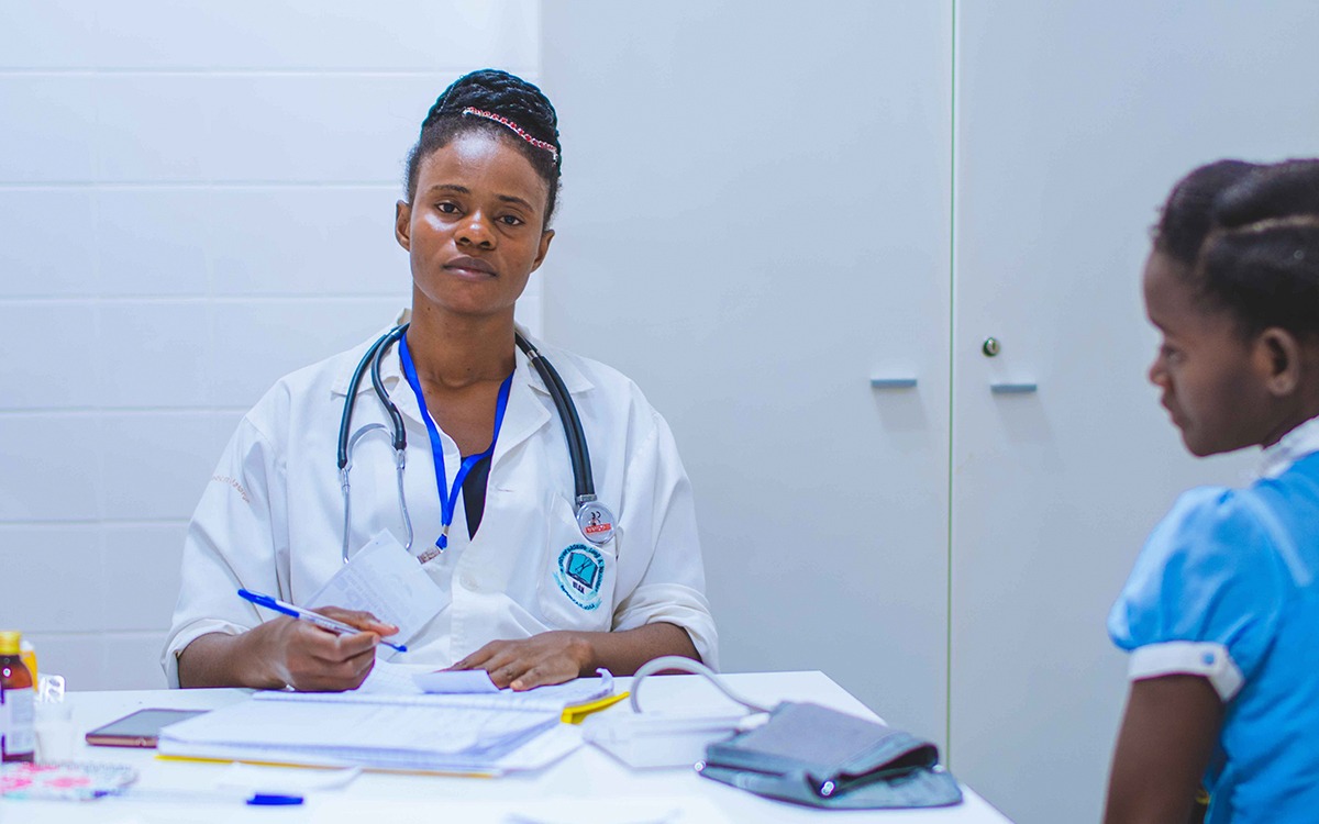A Black healthcare worker wearing a stethoscope and looking into the camera while writing on a notepad. There is a young patient in the foreground.