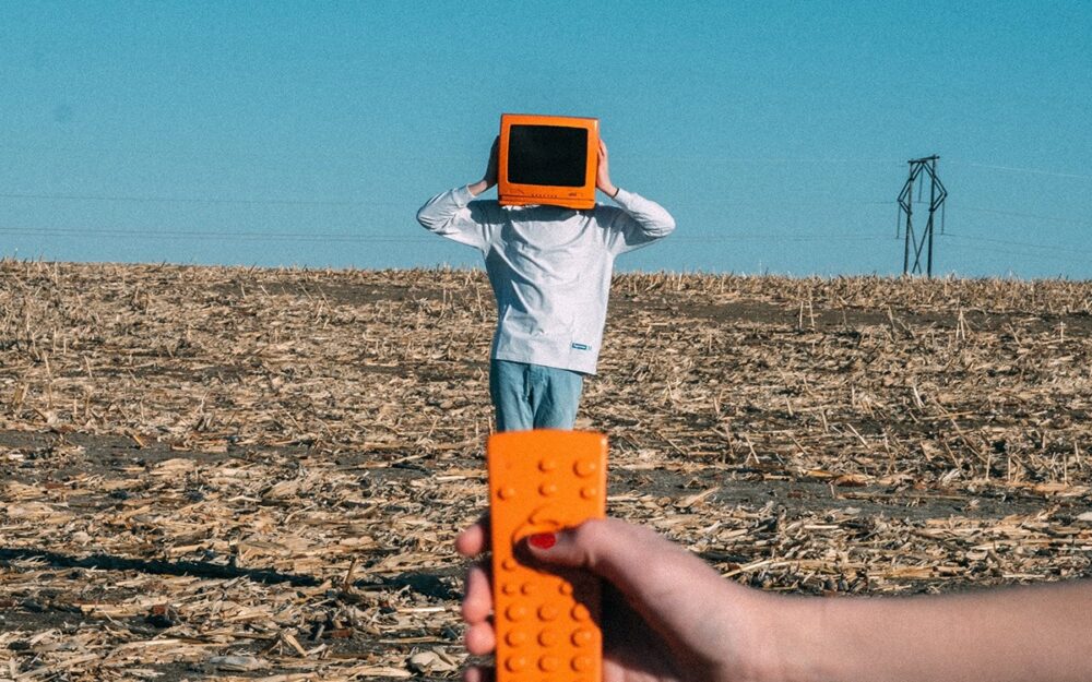  person with an orange television for a head standing in the middle of a barren field. There is a hand holding an orange remote in the foreground.