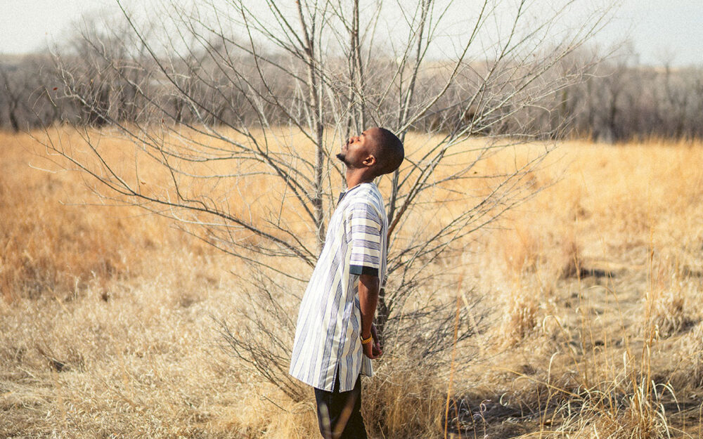 A Black man in a linen tunic standing in profile in front of a leafless tree in the middle of a field full of dead grasses. He is leaning back slightly with eyes closed.