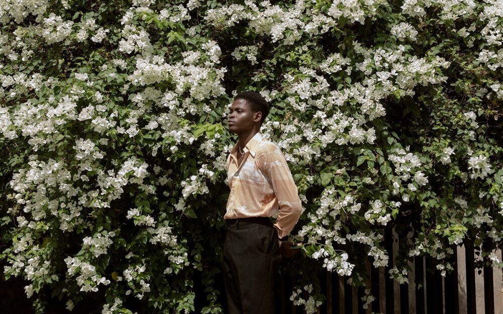 A Black man in a silk shirt standing in front of a blooming, flowery tree with white flowers.