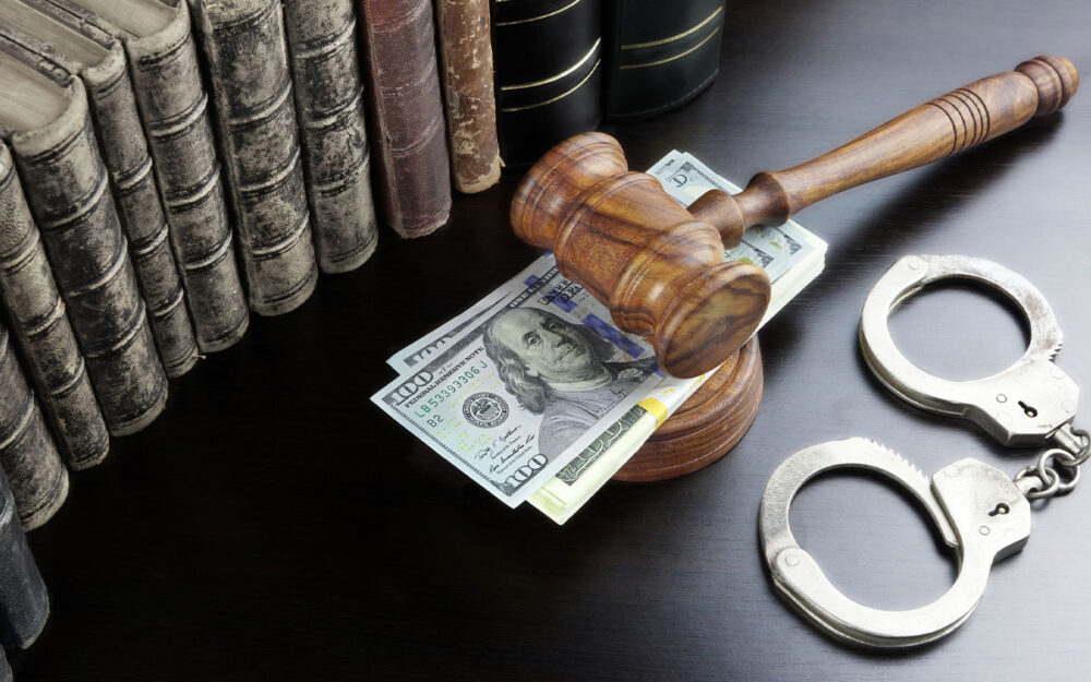 A judge’s gavel sitting on top of a pile of cash. There is a pair of handcuffs and law books surrounding the gavel.