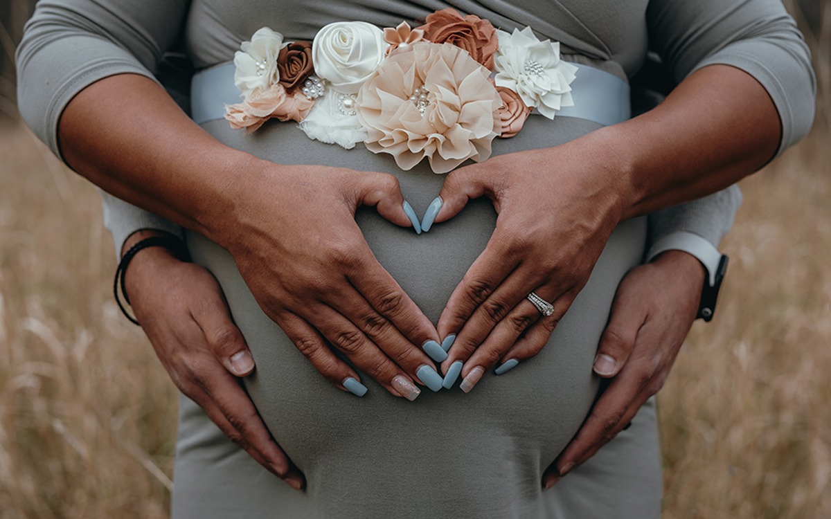 A close-up on the pregnant belly of a brown woman. Her hands make a heart over her belly, and her partner’s hand wrap around her hips from the back.