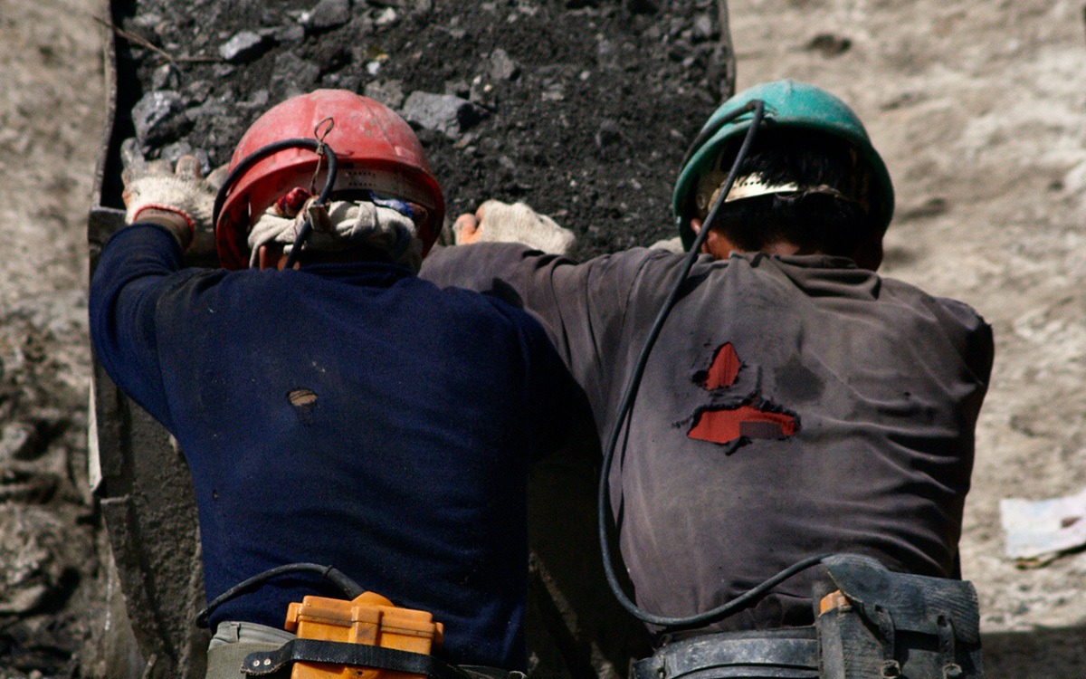Two coal miners with bowed heads and construction hats, pushing a loaded cart of coal