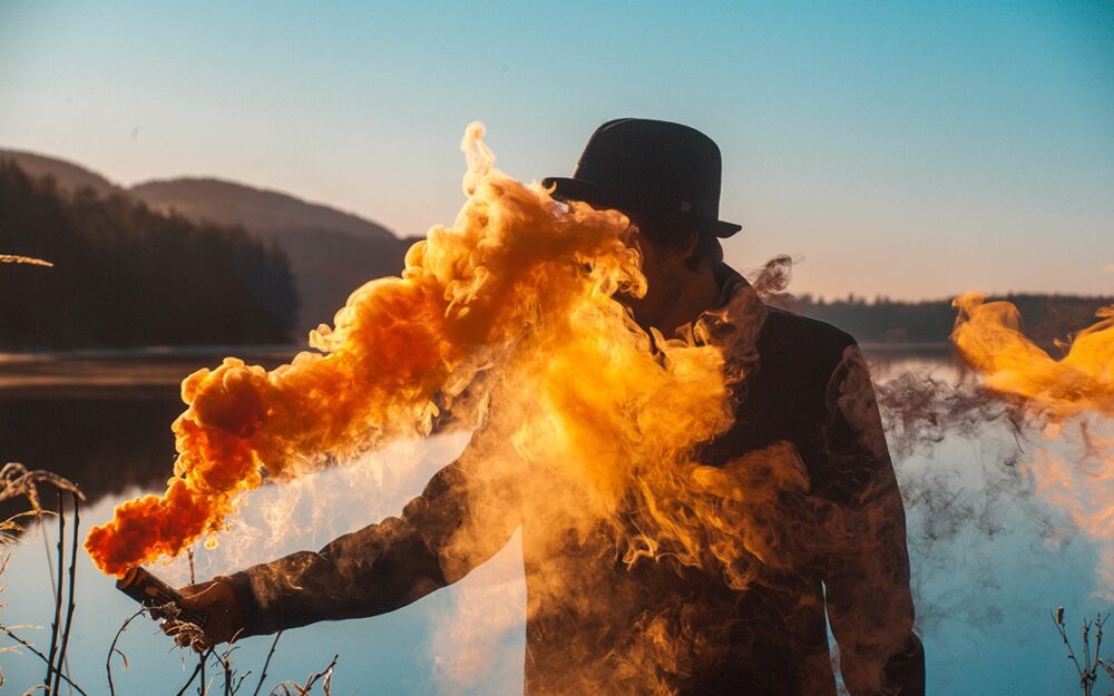 A man in a bowler hat, holding a smoke bomb releasing orange smoke. The smoke covers his face as he stands on the bank of a body of water.