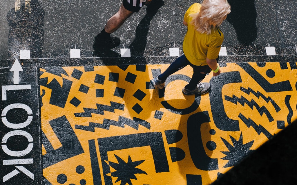 Bird’s eye view of people crossing a crosswalk painted with yellow and black designs. The word, “LOOK” is painted onto the ground.