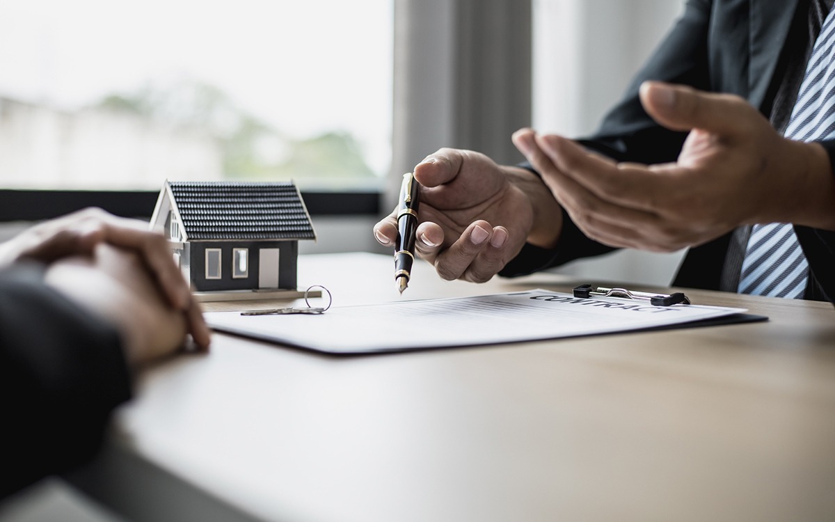 An employee explaining rent details to a tenant before signing the contract. The employee holds a pen and there are keys and a model house on the table.