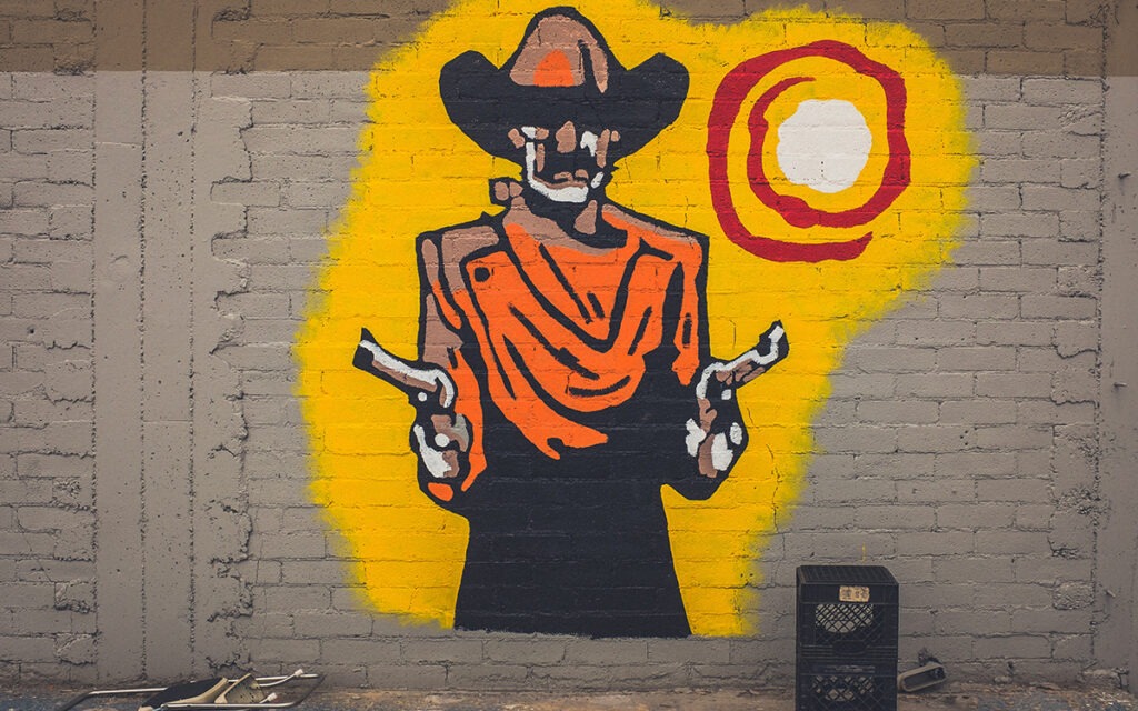 An alleyway mural of a mustachioed man wearing a cowboy hat and wielding two handguns. There is a sun blazing behind him.