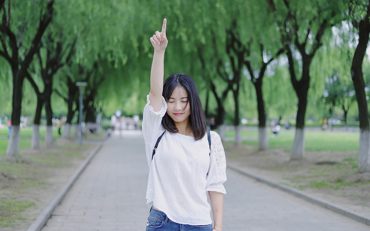 Young, asian woman standing on a tree-lined pathway and pointing up. She is smiling and closing her eyes.