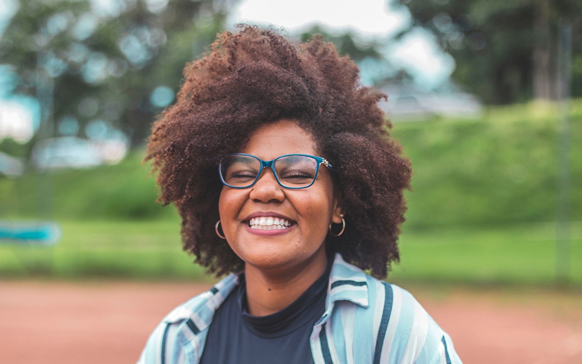 A Black woman with a brown, dense afro and blue glasses smiling widely and facing the camera.