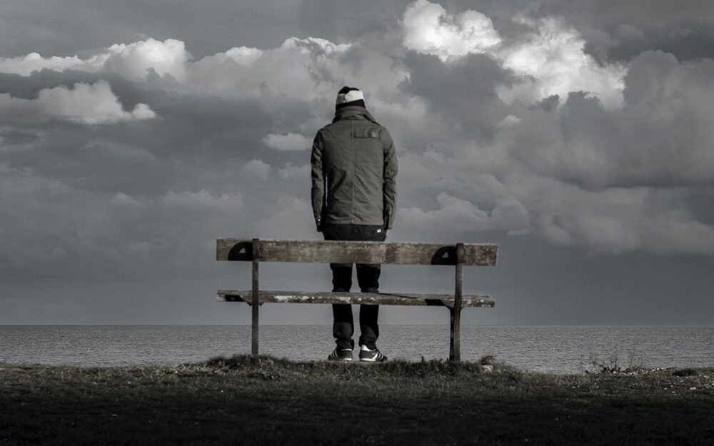 A desaturated image of a man standing in front of a bench over looking a body of water. His back is to the camera.