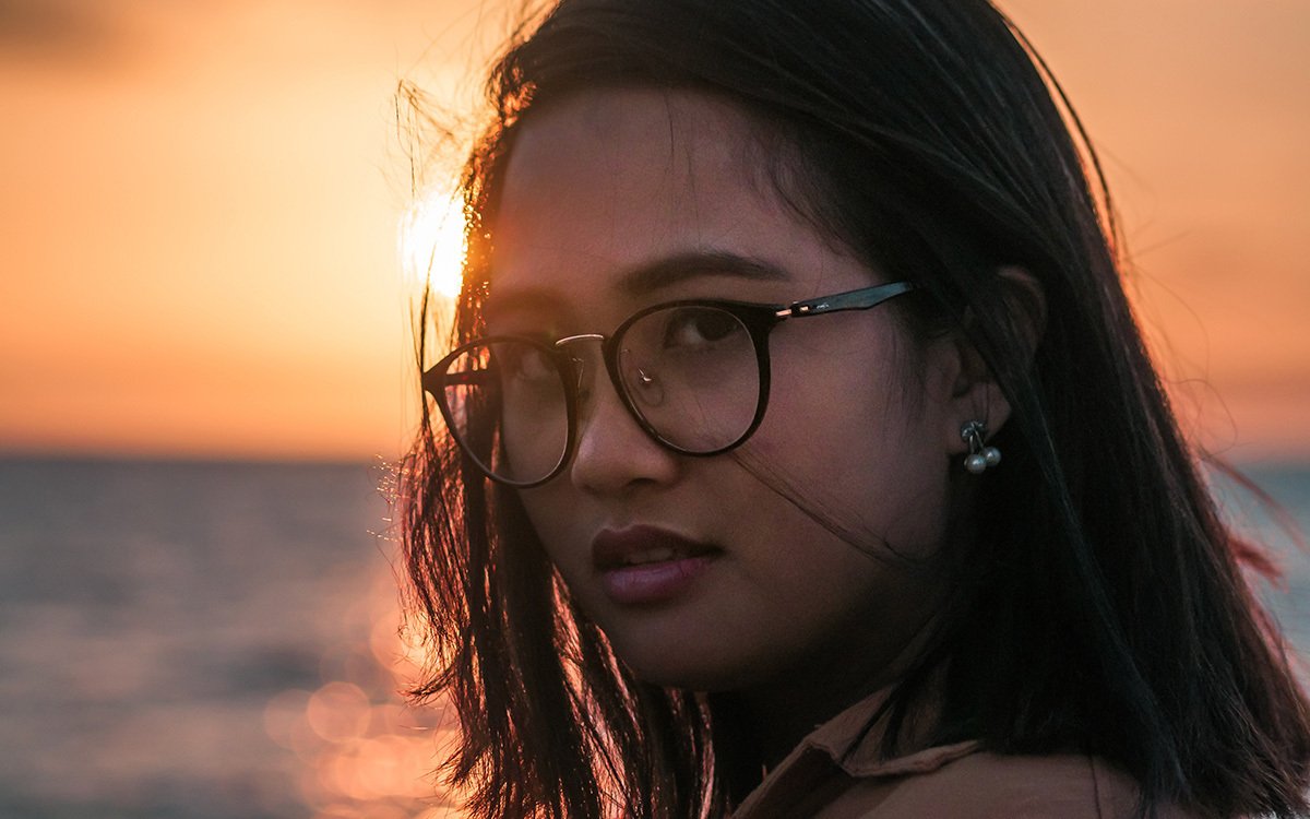 Close-up of an Asian woman in glasses, looking into the camera as the sun sets behind her.