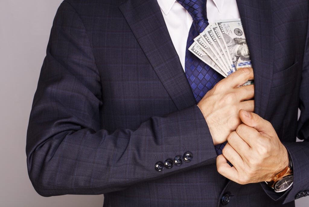 A man in a business suit stuffing a wad of 100 dollar bills into his interior suit jacket pocket discreetly.