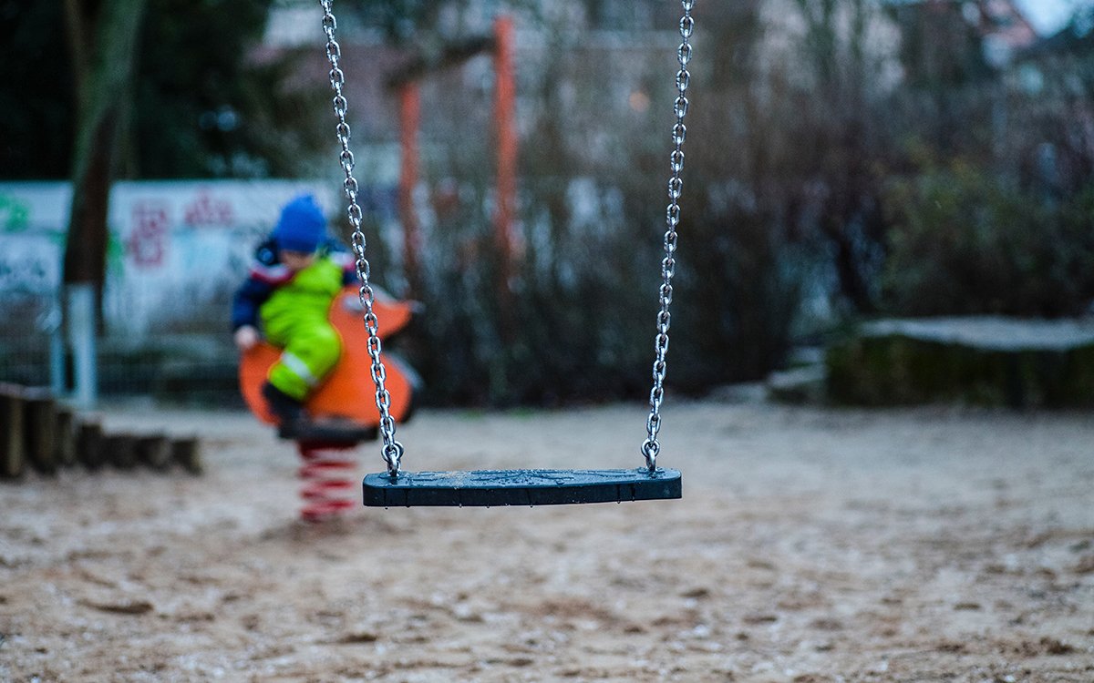 An empty swing at a playground. A child plays on a spring rider in the background.