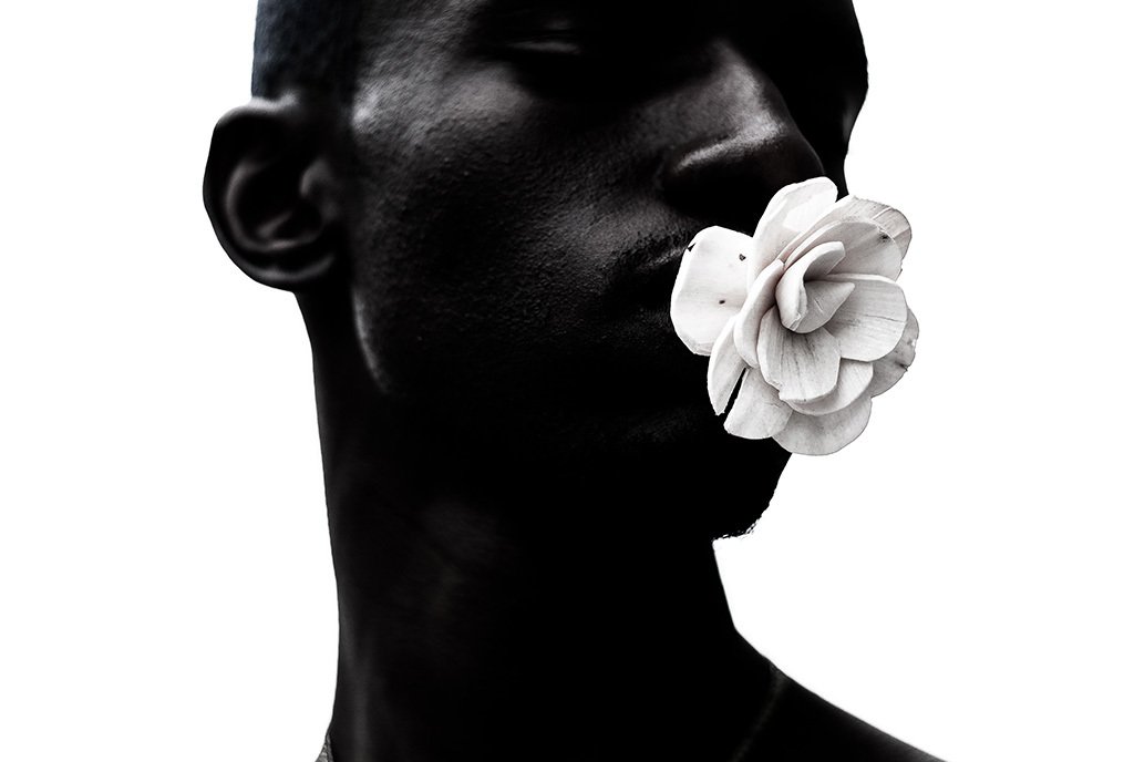 Black man with dark skin looks off into the distance as he holds a flower in his moths.