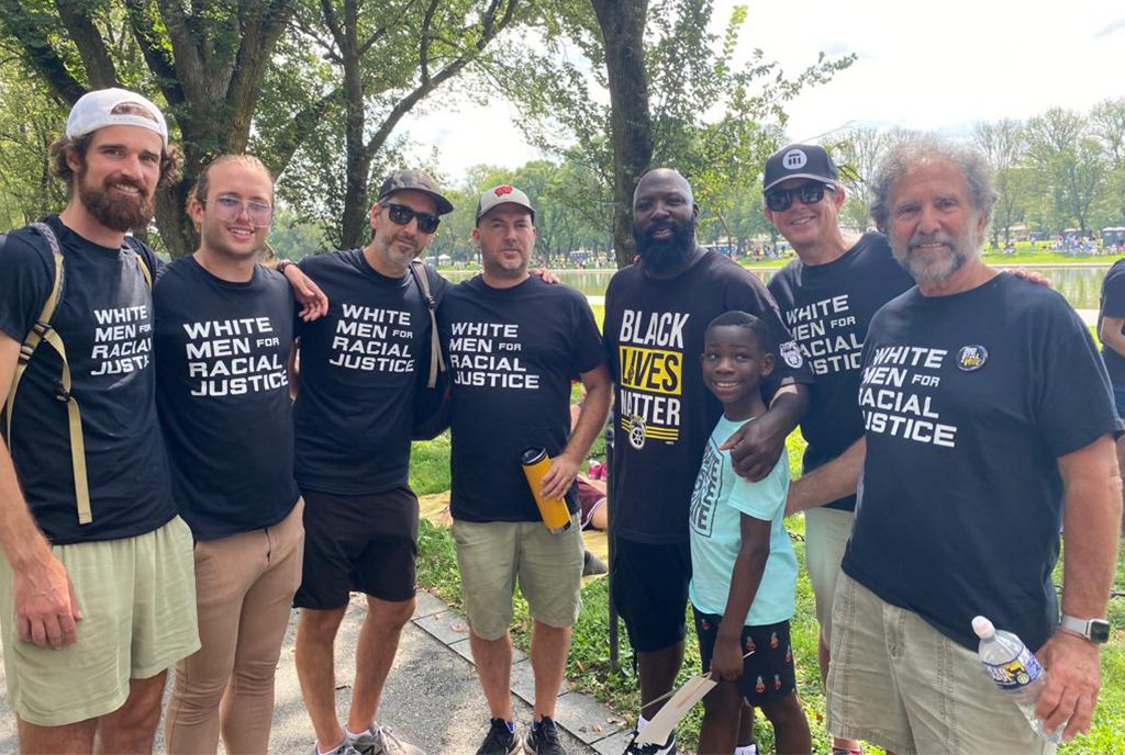 Group of several men, many of whom are White and wearing shirts that say, “White Men for Racial Justice”. There is a Black man and young boy in the picture.