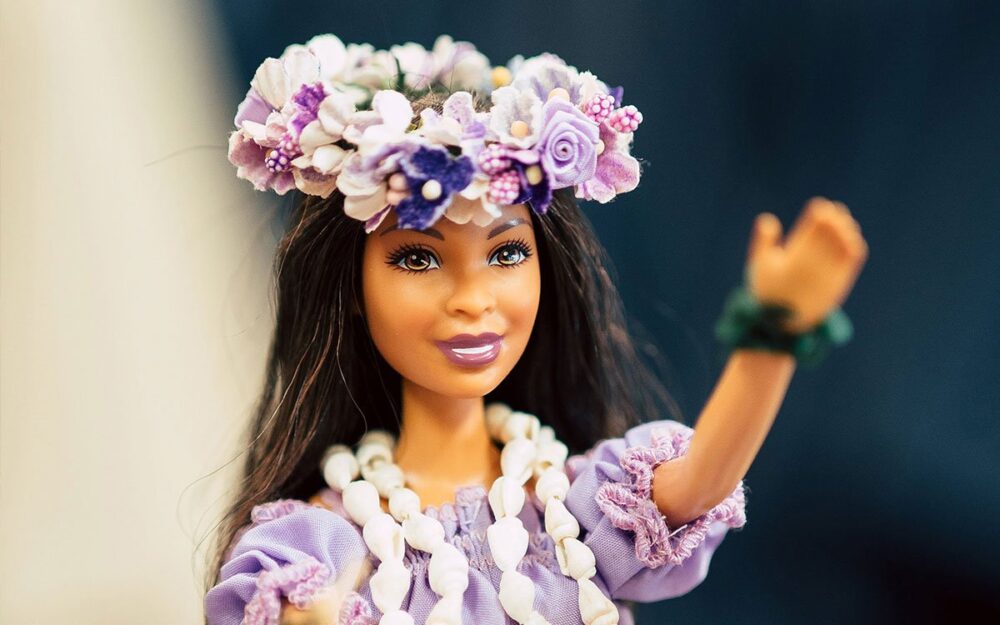 A brown-skin Barbie, wearing a cowrie shell necklace and traditional Hawaiian garb. She wears a lei on her head and extends her arm.