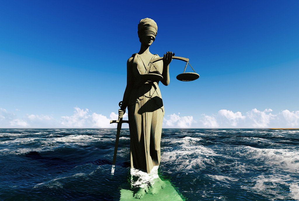 A statue of the Lady of justice, holding scales and standing in the ocean. The waves lap at her feet.