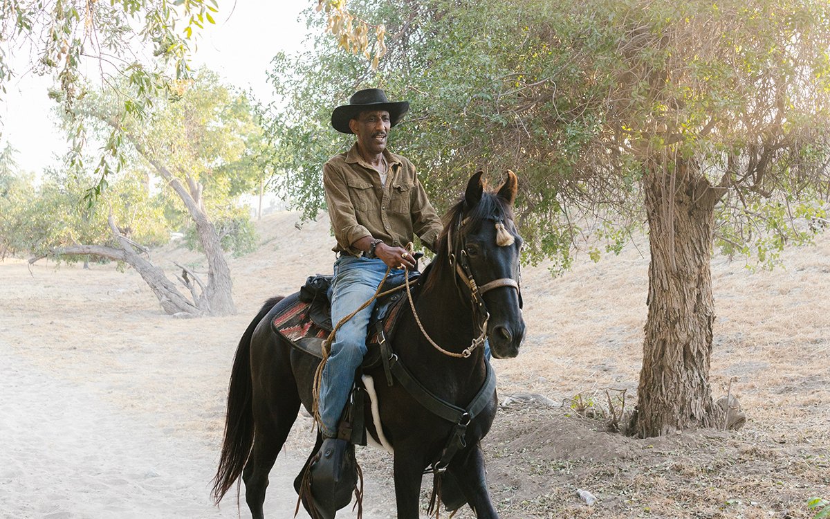 A Black man wearing a cowboy hat rides a Black horse down a country road