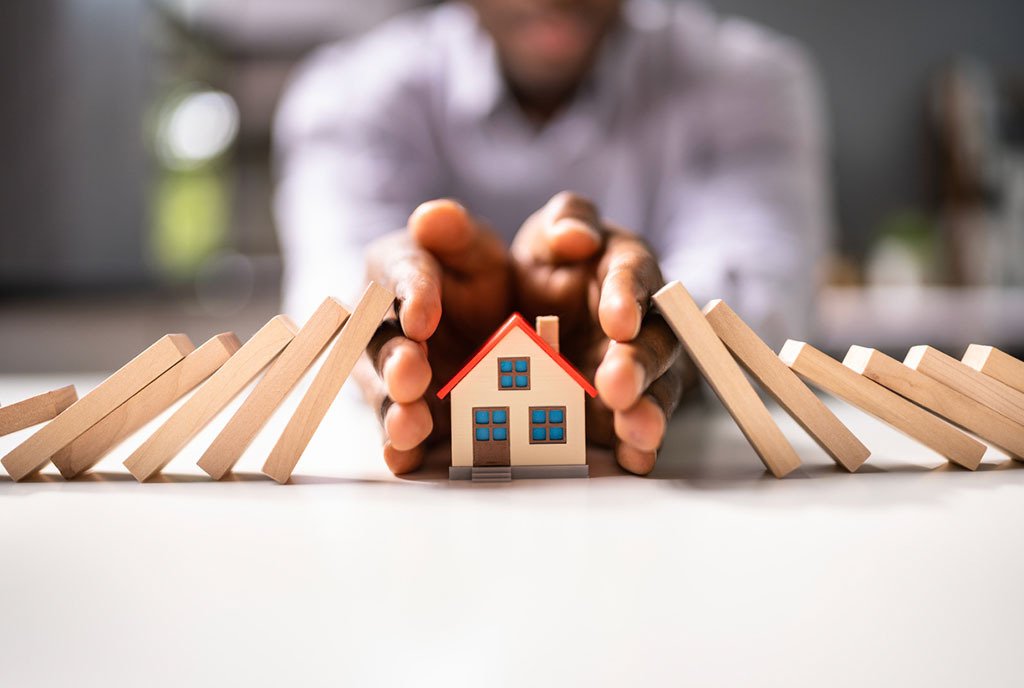 A man’s hands cradling and protecting a small toy house. There is a stack of toppled dominoes on both sides of his hands, from which he protects the house. To illustrate climate change’s affect on home insurance.