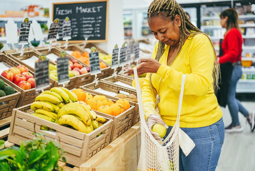 Black woman with braids in the produce section of a gentrified food co-op, putting an apple into a mesh bag.