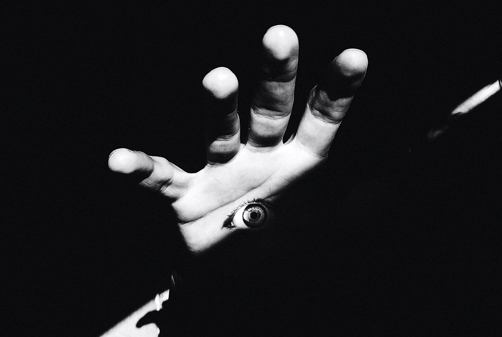A black and white photo of a hand in a dark shadow. There is a wide eyeball in the middle of the palm.