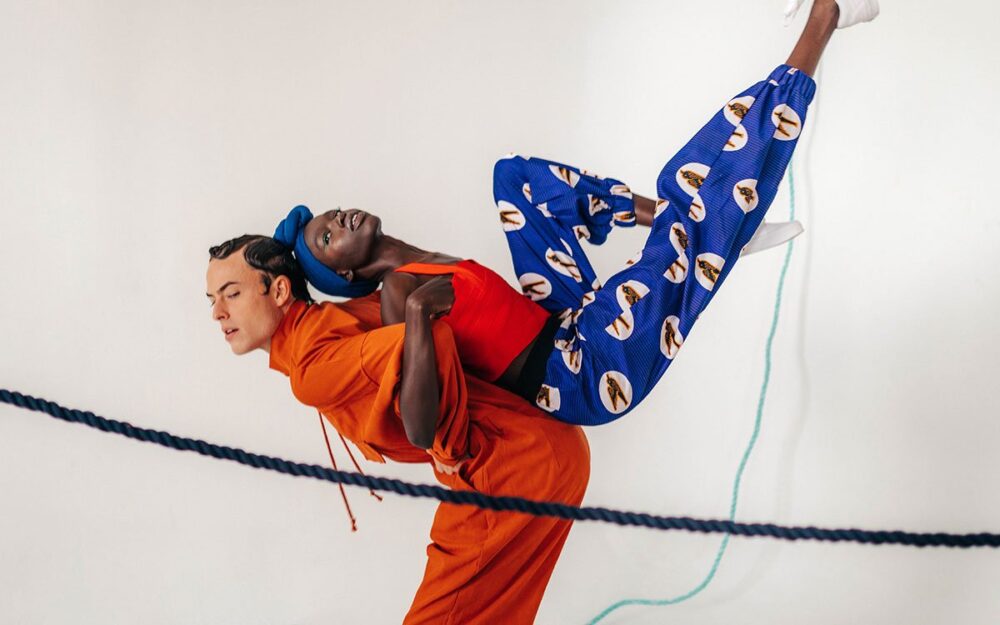A white man with coiffed hair interlocks arms with a black woman wearing a headscarf and patterned pants, and hoists her up on his back.