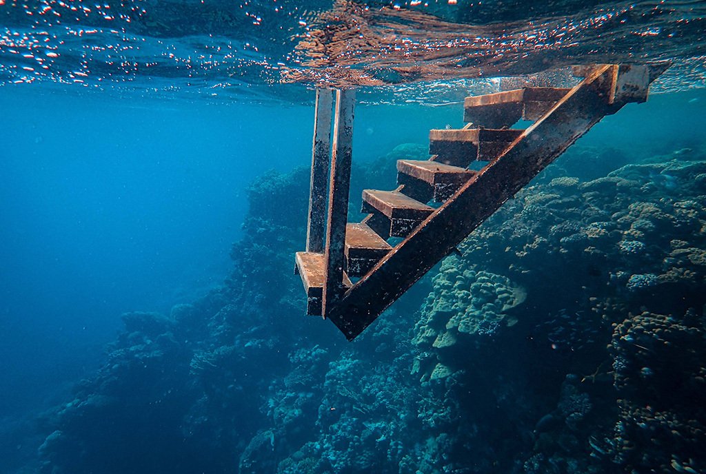  An underwater shot of a submerged wooden staircase raising out of the water. There is coral in the background.