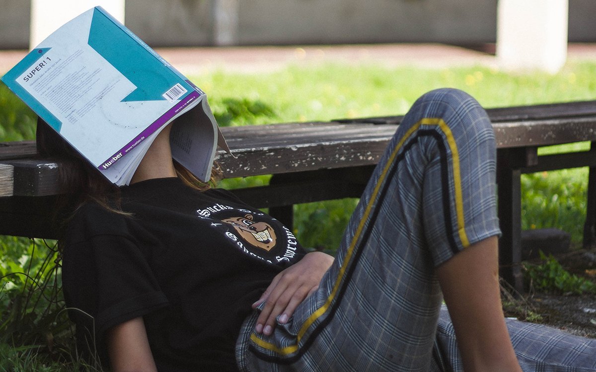 A college student looks exasperated sitting on the grass in front of a bench, with an opened book covering her face.