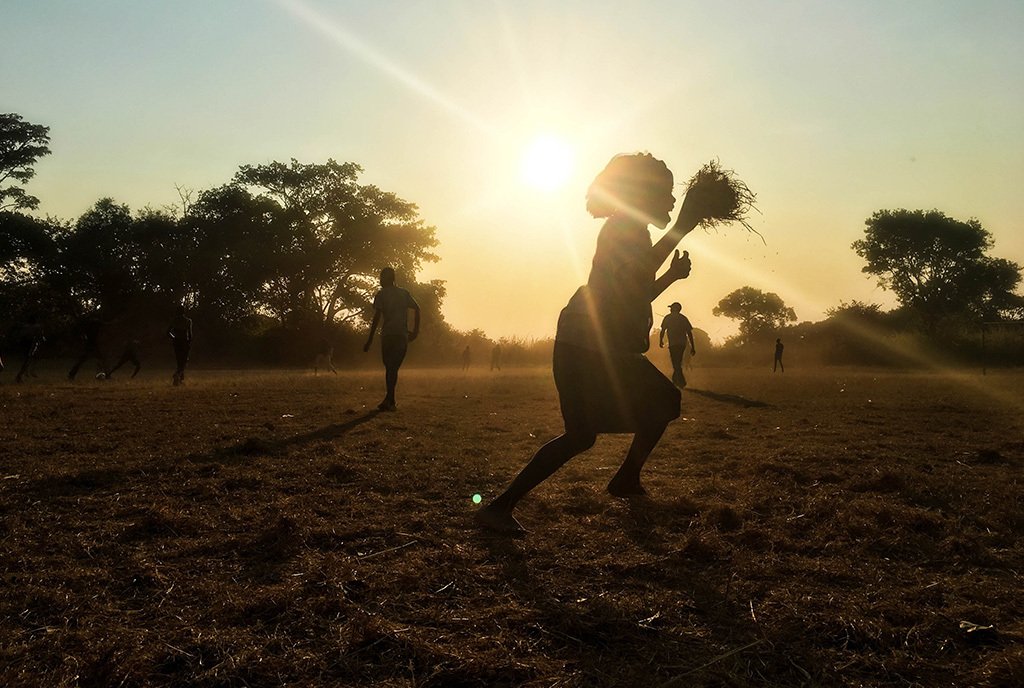 A young girl with grass in her hands runs across an empty field in Zambia. The sun sets in the background