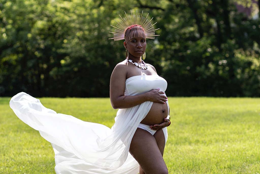 A pregnant Black woman standing in a grassy field, with her white gown flowing behind her. She is wearing an elaborate crown.