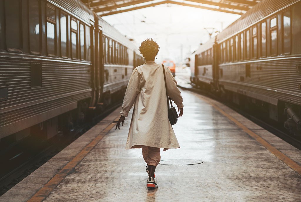 Black woman walking away from the camera, between two trains on a train platform.