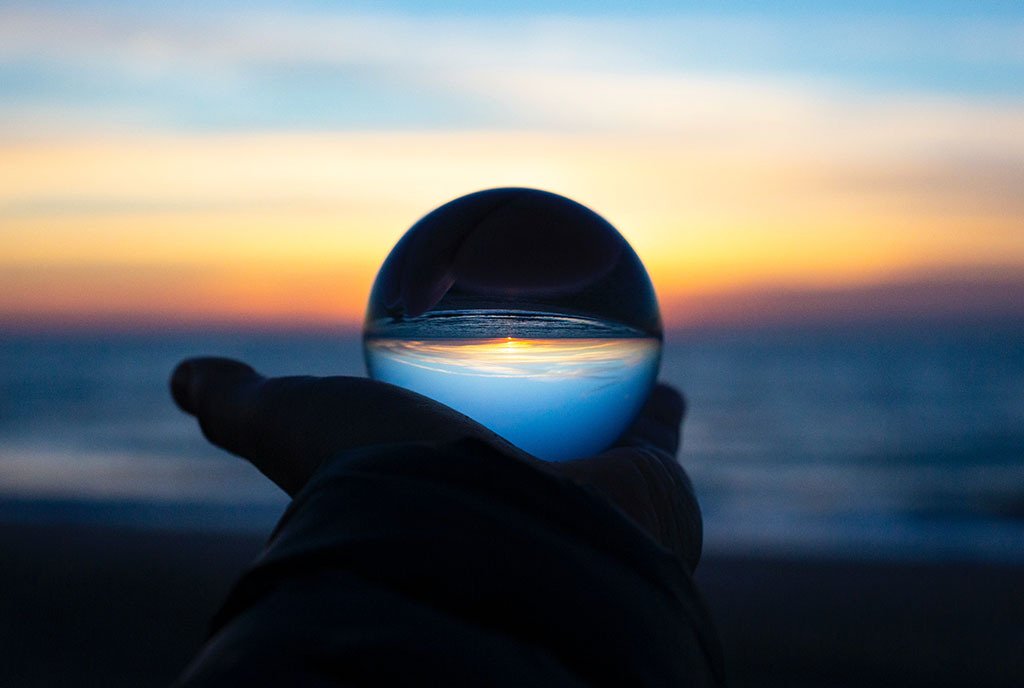 A hand holding up a glass orb that clarifies and intensifies the reflection of a sunrise in the distance