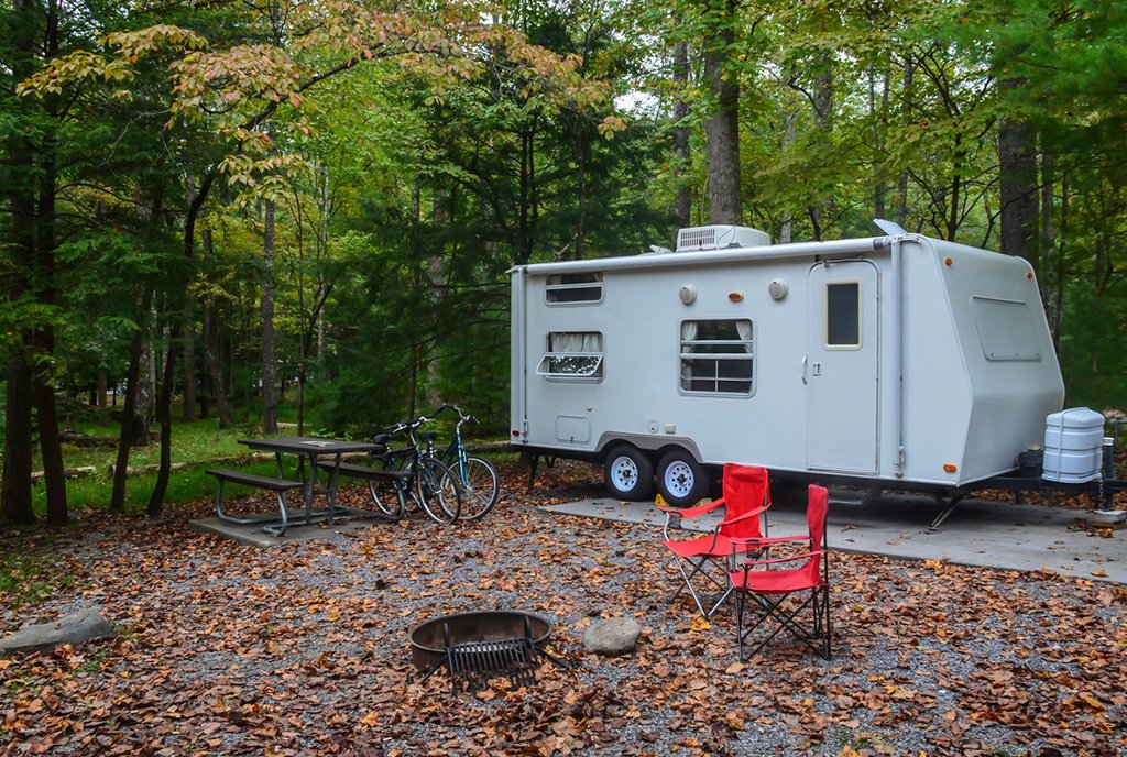 Camper trailer in wooded park camp site with chairs set up around fire pit, and bicycles leaning against picnic table.