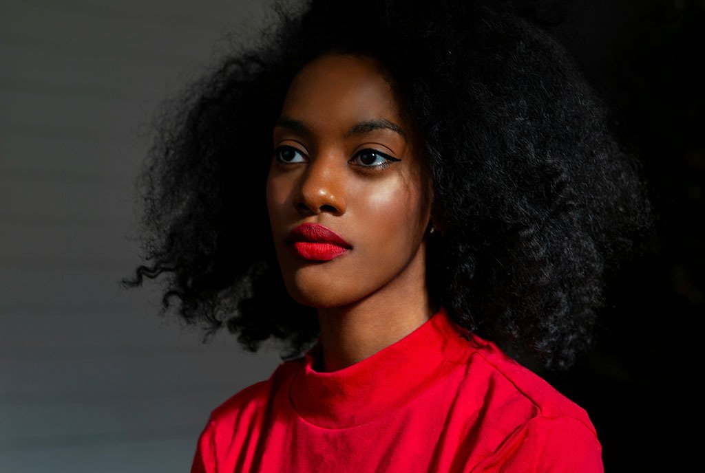A Black woman with natural hair, looking off into the distance, wearing blood-red lipstick and a red shirt.