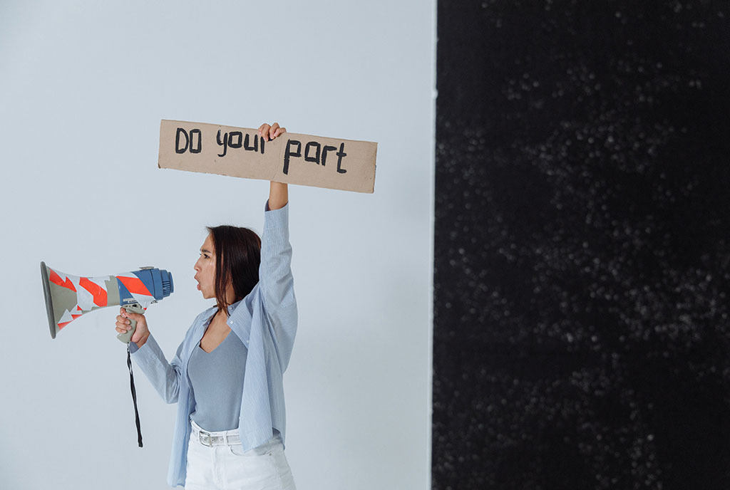 An Asian woman holding a megaphone and a cardboard sign that reads, “do your part”