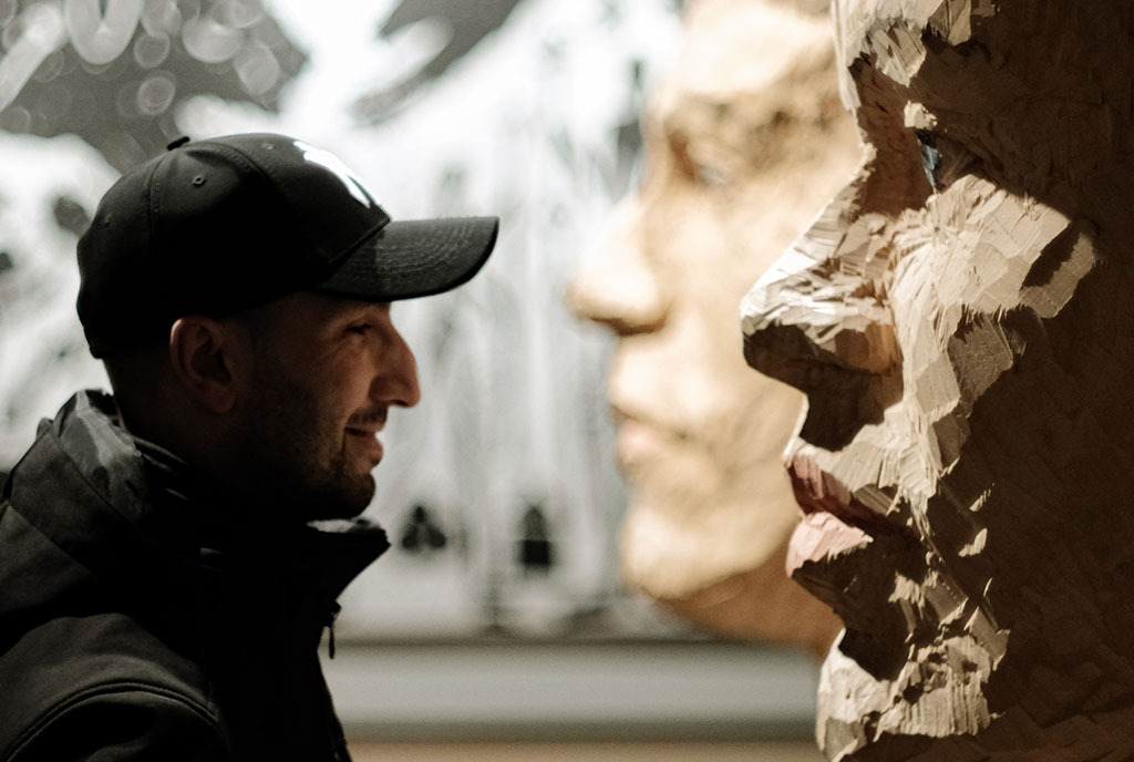 A man in a baseball cap facing a large, carved wooden face with a blank expression. To illustrate Anti-Immigrant Sentiment.