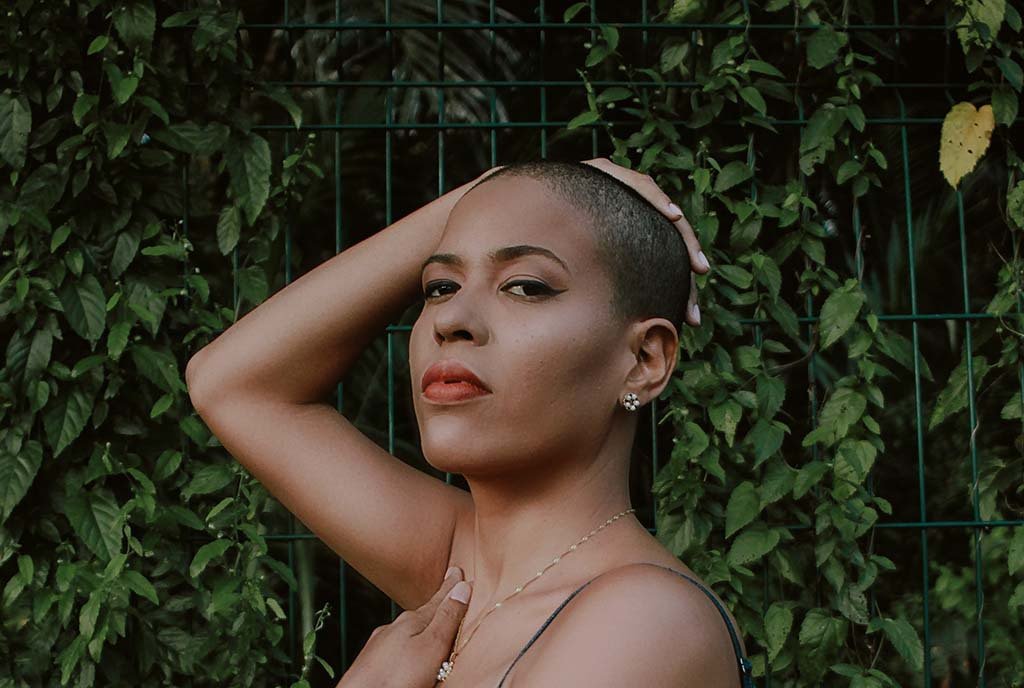 A Latino women with a buzzcut and red lips, looking into the camera and posing with her arm above her head.