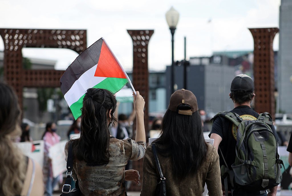 A group of protestors with their backs turned to the camera, marching and waving a Palestinian flag in the air.