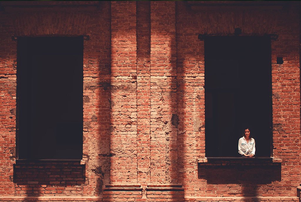 A pair of tall windows in a red brick wall. A Woman stands in one of the windows. To illustrate Abortion Access.