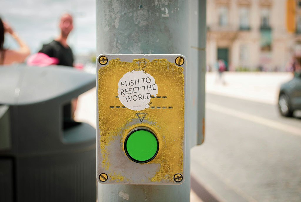 A sticker that reads “ push to reset the world”, pointing to a crosswalk button on a pole