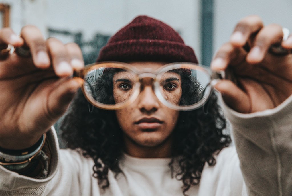 A man with long, wavy hair and a red beanie holding up glasses to the viewer, so as to make the image more clear.