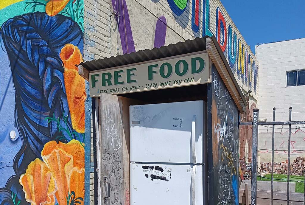 Community fridge in Oakland, California with the words “Free Food: Take What You Need. Leave What You Can” painted on the front. A colorful Mural reading “Build Unity” is in the background.