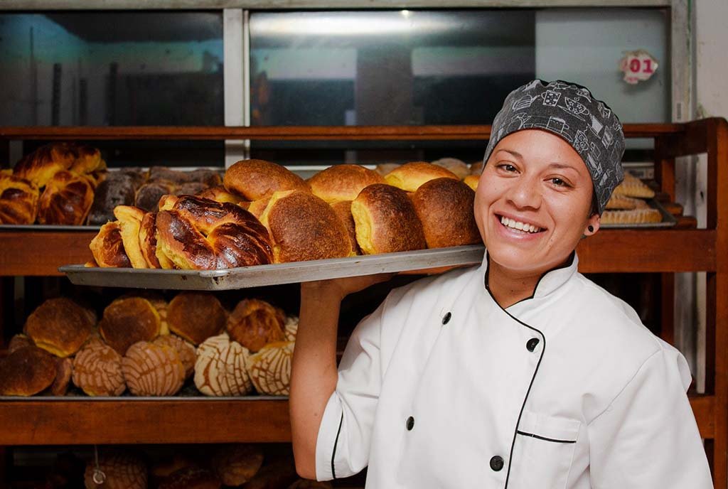 A latina baker at a local bakery, smiling and holding up a tray of baked bread