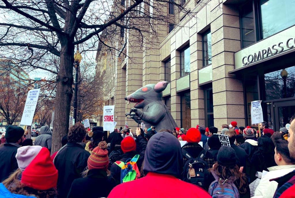 A crowd of people gathered on the Washington Post picket line. There is a inflatable Scabby the Rat among the crowd.