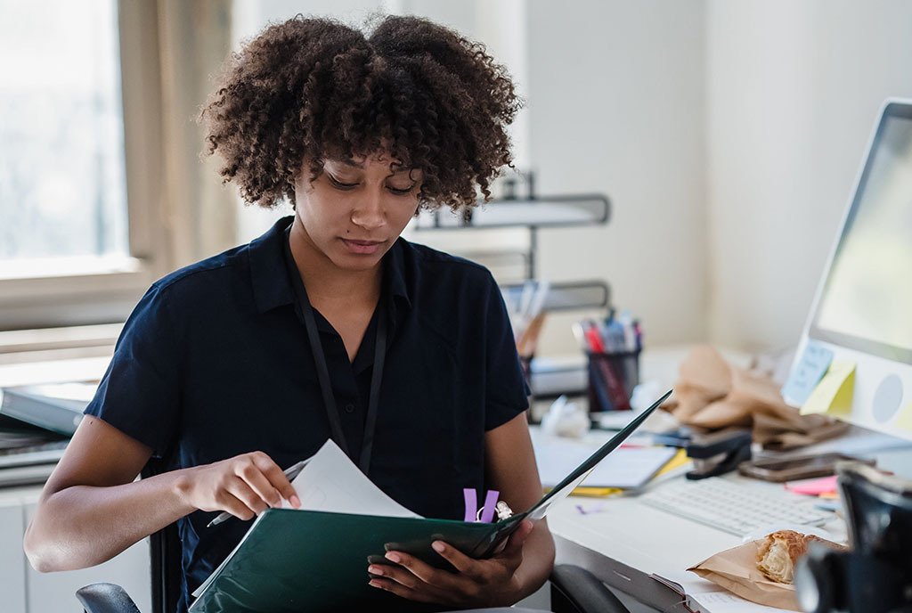 Black woman with curly hair sitting at an office desk reading documents in a folder.