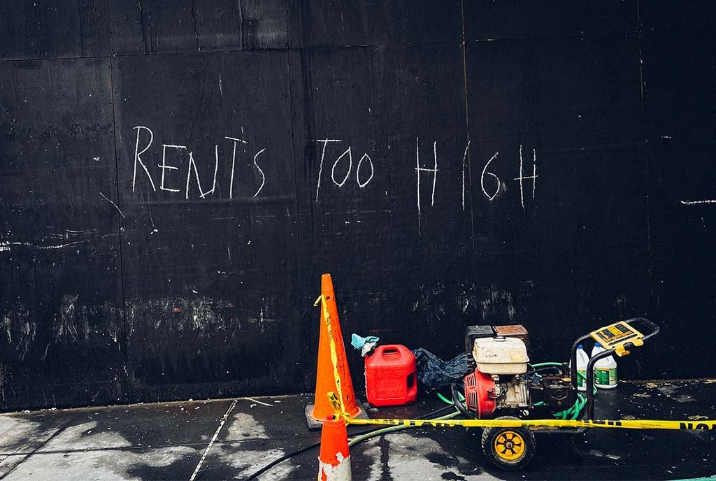 Photo of a chalked message reading, “Rent Is Too High” on a black plywood construction wall. There are construction materials in front of the wall