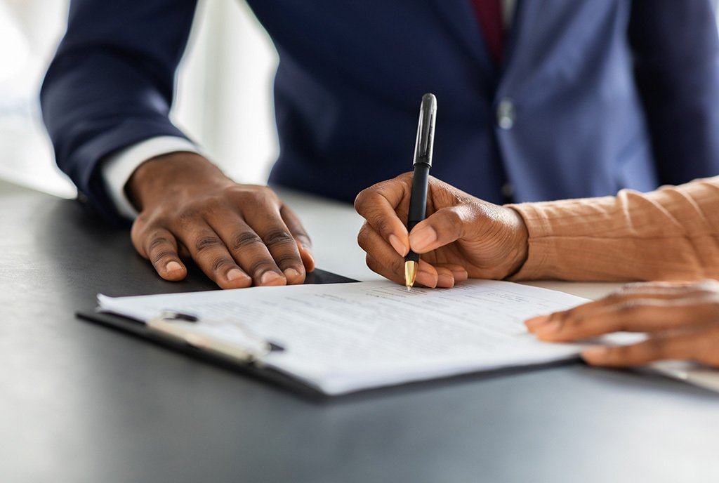 Close up shot of a Black female hand signing a contract with a pen. A Black man in a suit stands behind her.