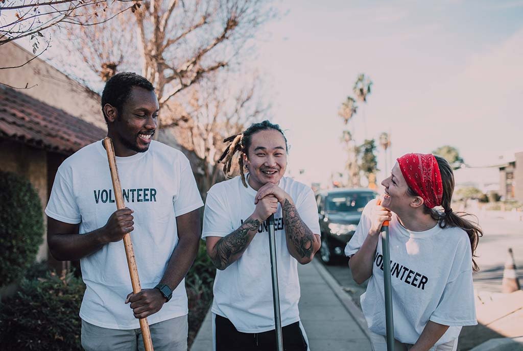 A Black man, an Asian man with dreadlocks, and a White woman in a red bandana, wearing “Volunteer” shirts and laughing together with brooms in hand.