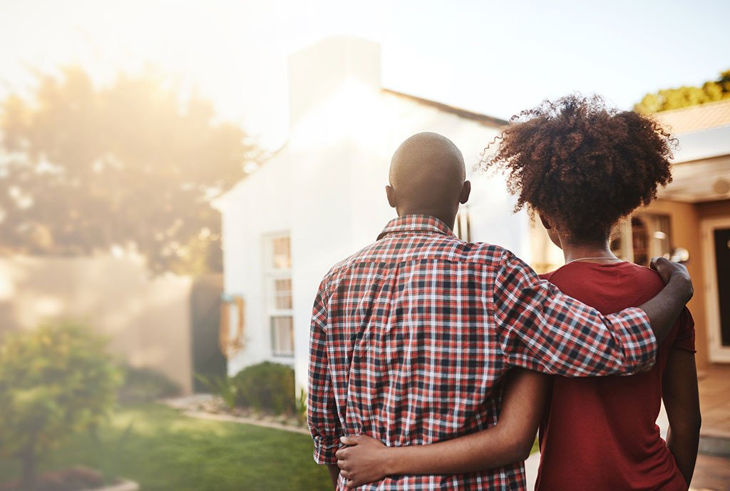A back view of a Black couple hugging each other in front of a house they have purchased.
